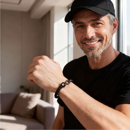 Lächelnder Mann mit Cap trägt das schwarze Flechtarmband mit Infinity-Anhänger am Handgelenk, Wohnzimmer im Hintergrund