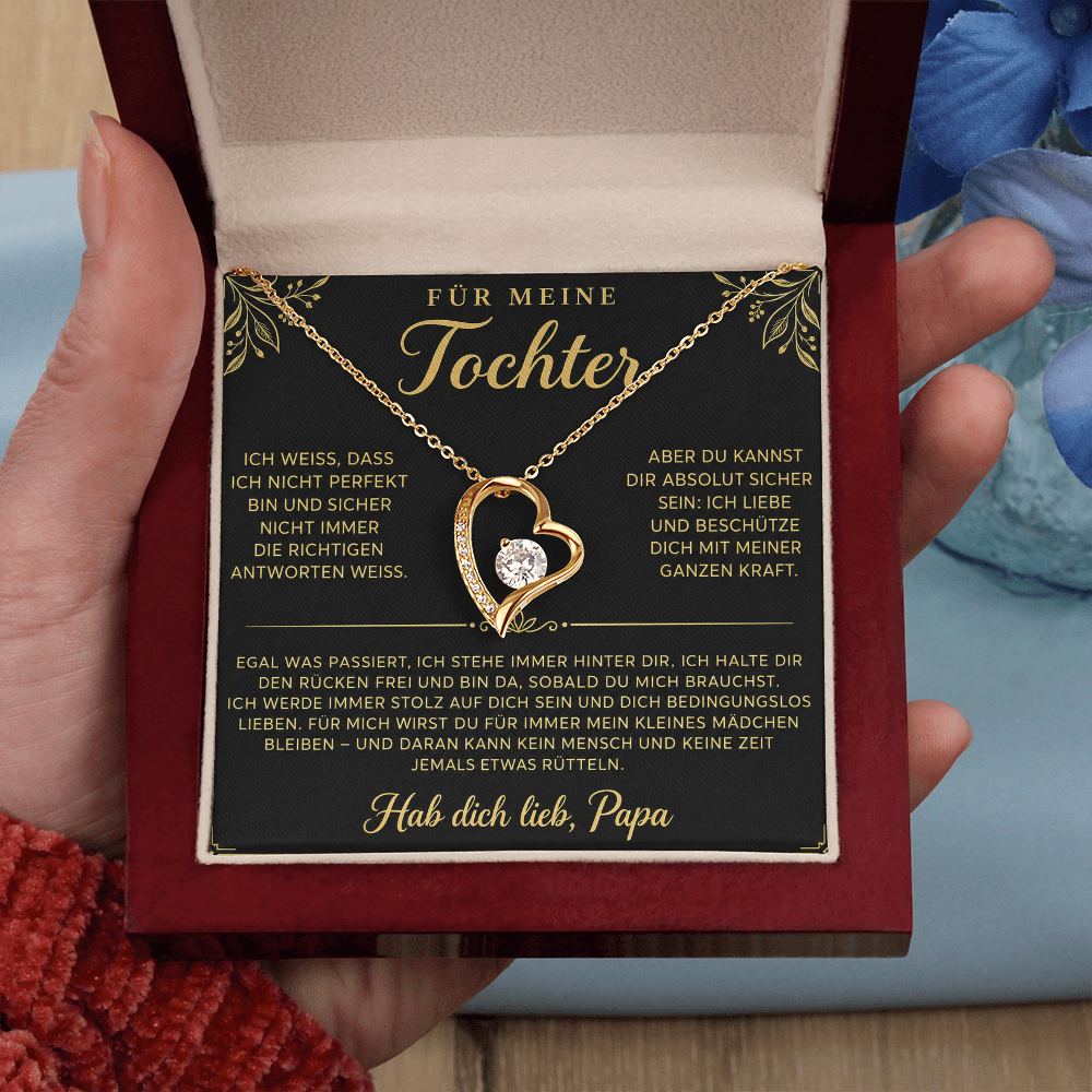 Hand hält die geöffnete Box mit goldenem Herzanhänger auf der Papa-Karte, blaue Blüten im Hintergrund