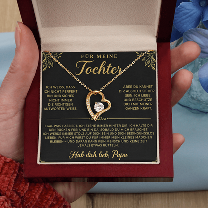 Hand hält die geöffnete Box mit goldenem Herzanhänger auf der Papa-Karte, blaue Blüten im Hintergrund