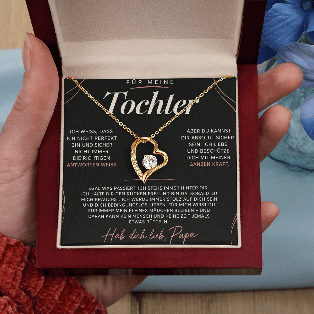 Hand hält die geöffnete Box mit goldenem Herzanhänger auf der roséfarbenen Papa-Karte, blaue Blüten im Hintergrund